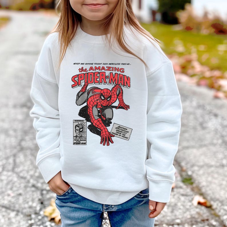 May include: White sweatshirt featuring a red and blue comic book image of Spider-Man. The text on the image reads "The Amazing Spider-Man".