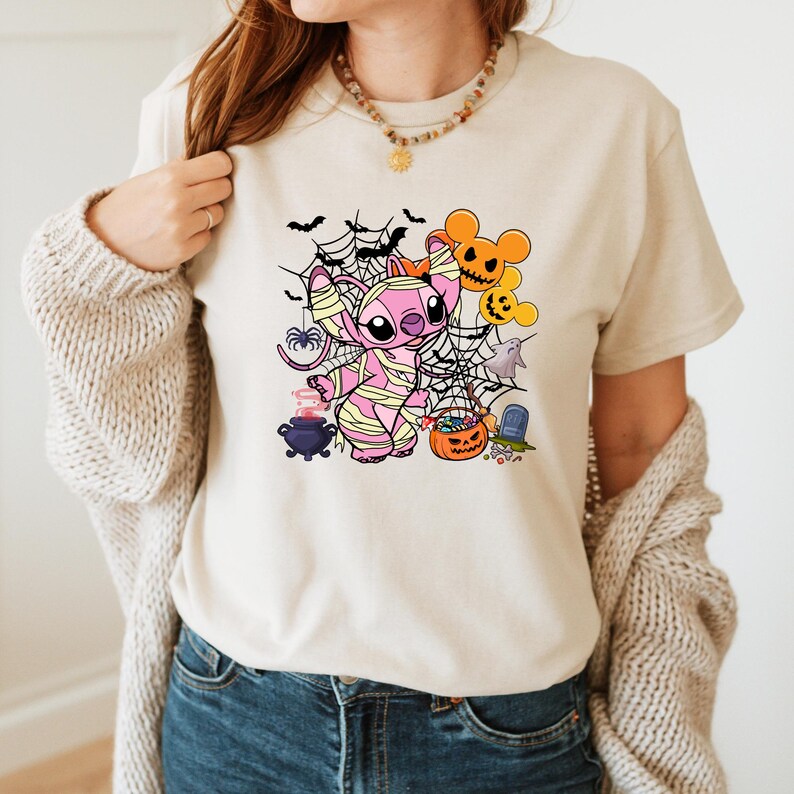 May include: Beige t-shirt featuring a Halloween-themed graphic. The design includes a pink mummy-wrapped character, spiderwebs, bats, and jack-o'-lanterns. A cauldron, ghost, and tombstone complete the spooky scene. The shirt is a casual, festive apparel item.