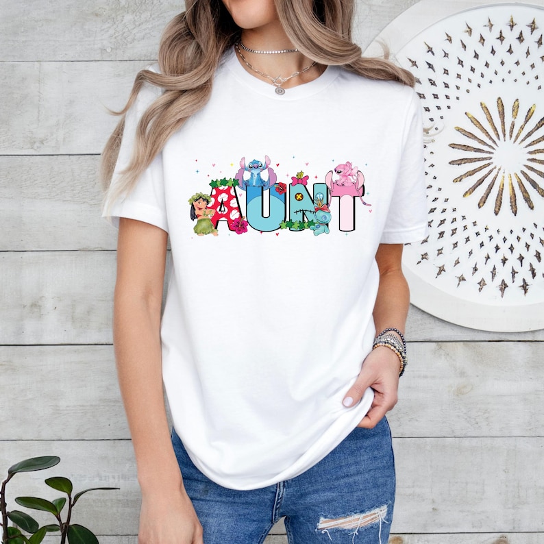 Puede incluir: Camiseta blanca con un gr&aacute;fico colorido de personajes de dibujos animados que deletrean "AUNT" con flores y corazones.