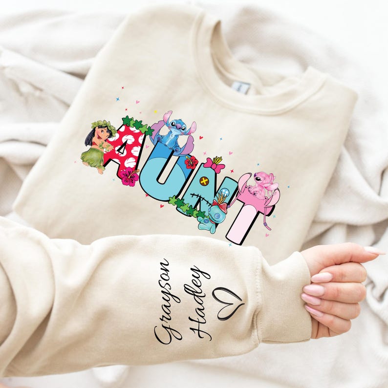 May include: A beige sweatshirt with a colorful "AUNT" graphic featuring Stitch and Lilo characters. The text "Grayson Hadley" is written on the sleeve with a heart.