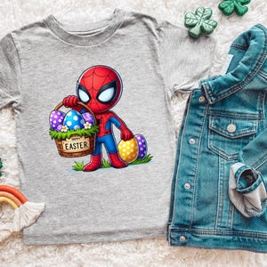 Puede incluir: Una camiseta gris con un personaje de dibujos animados de Spider-Man sosteniendo una cesta de huevos de Pascua. La cesta tiene el texto "HAPPY EASTER". Tambi&eacute;n se ve una chaqueta vaquera y decoraciones de tr&eacute;bol.