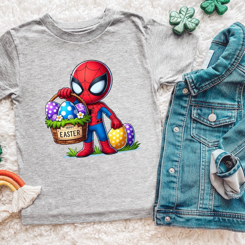 Puede incluir: Una camiseta gris con un personaje de dibujos animados de Spider-Man sosteniendo una cesta de huevos de Pascua. La cesta tiene el texto "HAPPY EASTER". Tambi&eacute;n se ve una chaqueta vaquera y decoraciones de tr&eacute;bol.