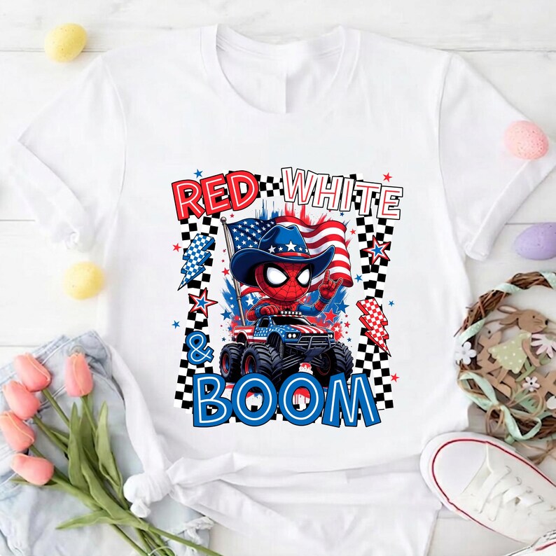 May include: White t-shirt featuring a cartoon Spider-Man character in a cowboy hat driving a monster truck. The design includes the text "RED WHITE & BOOM" with American flag and star graphics. The shirt is decorated with red, white, and blue colors.