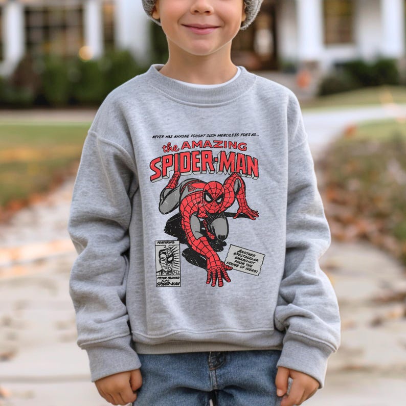 May include: A grey sweatshirt with a vintage comic book design featuring Spider-Man. The text on the shirt reads "Never has anyone fought such merciless foes as... The Amazing Spider-Man".