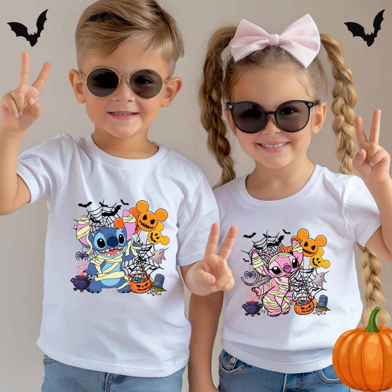 May include: Two children wearing white t-shirts with Halloween-themed cartoon graphics. The designs feature a blue and pink character wrapped in bandages, surrounded by spiderwebs, bats, and pumpkins. Both children are wearing sunglasses and making peace signs.