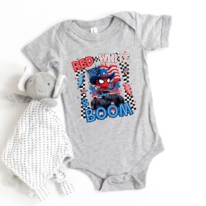 May include: A gray baby onesie with a patriotic graphic featuring a Spider-Man-like character in a cowboy hat on a monster truck, with the text "RED WHITE & BOOM". A stuffed elephant toy is next to the onesie.
