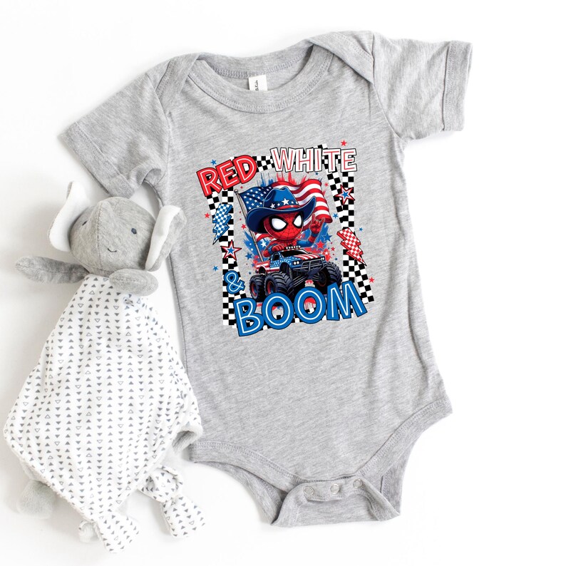May include: A gray baby onesie with a patriotic graphic featuring a Spider-Man-like character in a cowboy hat on a monster truck, with the text "RED WHITE & BOOM". A stuffed elephant toy is next to the onesie.