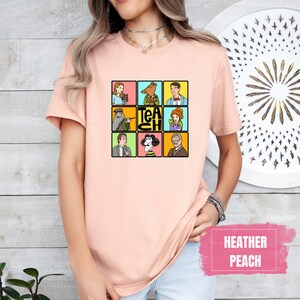 May include: Heather peach-colored t-shirt featuring a grid of cartoon characters with the word "TEACH" in the center. The design includes various figures in colorful squares. The shirt is a soft, casual style.