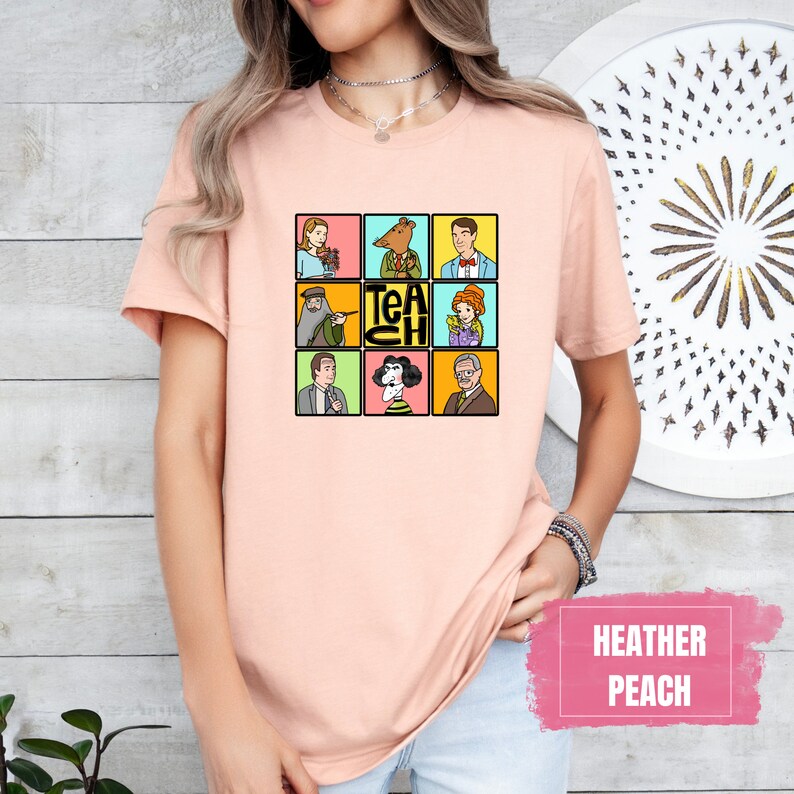 May include: Heather peach-colored t-shirt featuring a grid of cartoon characters with the word "TEACH" in the center. The design includes various figures in colorful squares. The shirt is a soft, casual style.