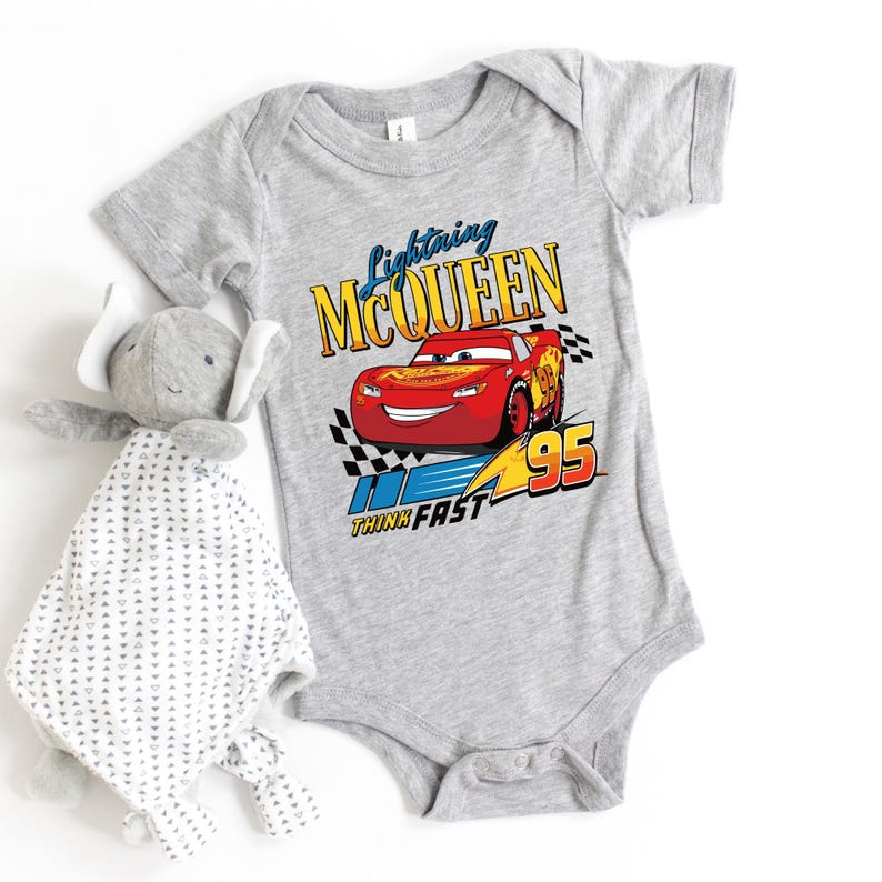 May include: A gray baby onesie featuring Lightning McQueen from the Cars movie. The design includes the text "Lightning McQueen" above a red race car, with "95" and "Think Fast" below. A gray elephant plush toy is next to the onesie.