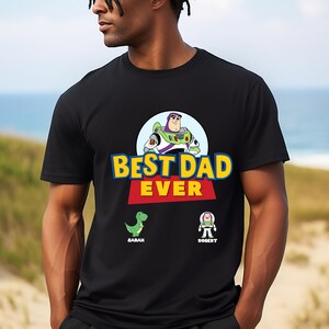 K&ouml;nnte beinhalten: Schwarzes T-Shirt mit einem Motiv von Buzz Lightyear aus Toy Story. Der Text "Best Dad Ever" ist in Rot und Gelb gedruckt. Unter dem Text befinden sich zwei kleinere Grafiken von Figuren aus Toy Story, mit den Namen "Sarah" und "Robert" darunter.