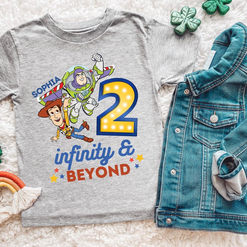 K&ouml;nnte beinhalten: Ein graues T-Shirt mit einer bunten Grafik von Buzz Lightyear und Woody aus Toy Story, der Zahl 2 und dem Schriftzug "infinity & beyond". Eine Jeansjacke liegt neben dem Shirt.