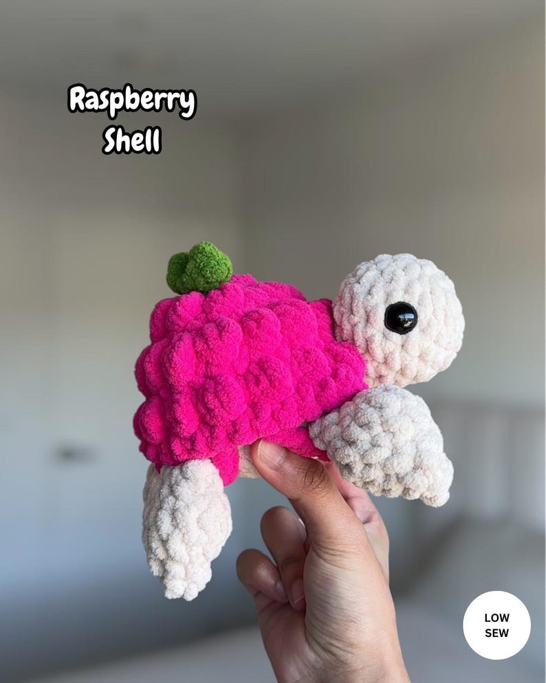May include: A crochet turtle plush with a bright pink raspberry-shaped shell, a small green leaf on top, and a cream-colored head and limbs. The turtle has black plastic safety eyes. Text on the image reads Raspberry Shell and LOW SEW.