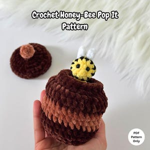 Könnte beinhalten: Ein handgefertigtes Häkel-Pop-It-Spielzeug in Form einer Honigbiene in Braun- und Gelbtönen. Das Spielzeug zeigt eine Biene, die aus einem braunen Behälter kommt. Der Text "Crochet Honey-Bee Pop It Pattern" steht oben.