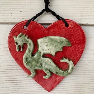 May include: A red heart-shaped ceramic ornament with a green dragon design. The ornament has a black cord for hanging.
