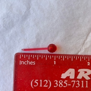May include: A small, red, faceted Christmas light bulb with a clear stem, approximately 1 inch long, rests on a red ruler. The ruler displays the word "Inches" and a phone number.