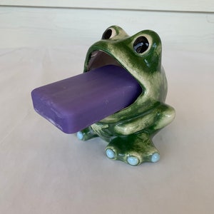 May include: A green ceramic frog soap dish with a purple bar of soap in its mouth. The frog has a white mouth and black eyes.