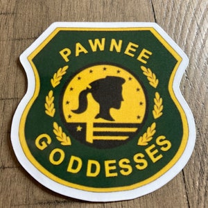 May include: A sticker with a green and gold shield design. The sticker features the words "PAWNEE GODDESSES" in white and gold lettering. A silhouette of a woman with a ponytail is in the center.
