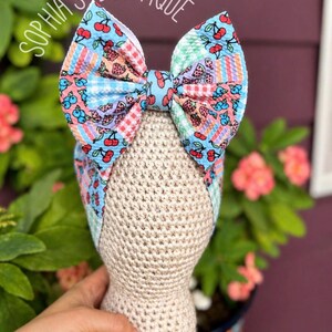 May include: A colorful headband with a large bow featuring a fruit pattern of cherries, strawberries, and watermelon. The headband is displayed on a mannequin head. The text "SOPHIA'S BOWTIQUE" is arched above the bow.