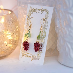 May include: A pair of gold dangle earrings with red berry clusters and green leaves. The earrings are displayed on a white card with a gold border.