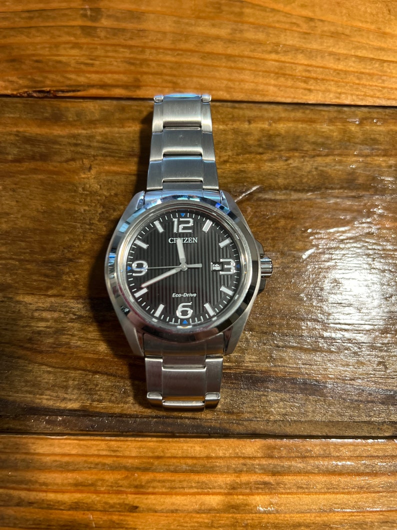 Puede incluir: Un reloj Citizen Eco-Drive plateado con esfera negra y agujas e indicadores plateados. El reloj tiene una correa de acero inoxidable y una ventana de fecha. El reloj se muestra sobre una superficie de madera.