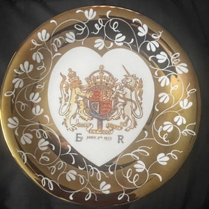 May include: A decorative plate with a gold rim and a white heart-shaped center. The center features a royal coat of arms and the text "June 2nd 1953". The plate is adorned with white floral designs.