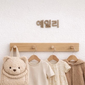 May include: A beige teddy bear backpack hangs on a wooden rack with a short-sleeved shirt, floral dress, and a fuzzy jacket. A small wooden table holds toys, and a stuffed bunny sits near a woven basket. The Korean text "에일리" is above the rack.