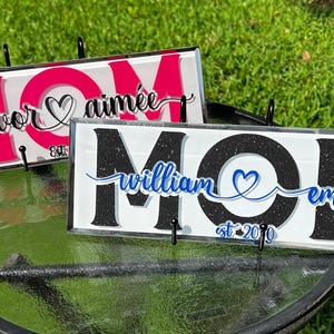 May include: Two decorative glass signs with the word "MOM" in large black letters. The top sign is pink with white lettering and says "Trevor & Aimee est 2019". The bottom sign is black with white lettering and says "William & Emma est 2019".