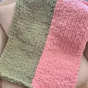 May include: A soft, chunky knit blanket in two colors: sage green and light pink. The blanket is folded over a neutral-colored chair, showcasing the texture and color contrast. The thick yarn creates a cozy, inviting look.