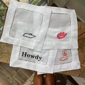 Peut inclure: Quatre serviettes en lin blanc avec des motifs brod&eacute;s. Une serviette repr&eacute;sente le contour du Tennessee avec "TN" &agrave; l'int&eacute;rieur. Une autre serviette a un chapeau de cowboy rose. Une troisi&egrave;me serviette a le mot "Howdy" brod&eacute; dessus. La derni&egrave;re serviette a une botte de cowboy rose.