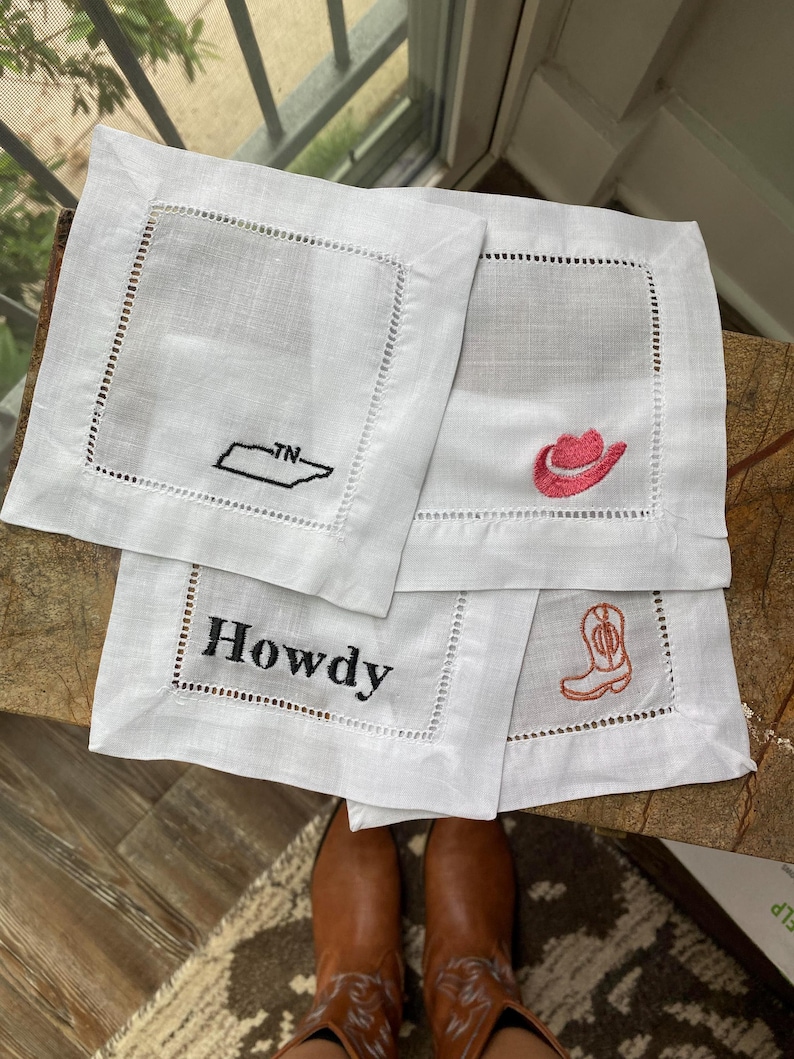 Peut inclure: Quatre serviettes en lin blanc avec des motifs brod&eacute;s. Une serviette repr&eacute;sente le contour du Tennessee avec "TN" &agrave; l'int&eacute;rieur. Une autre serviette a un chapeau de cowboy rose. Une troisi&egrave;me serviette a le mot "Howdy" brod&eacute; dessus. La derni&egrave;re serviette a une botte de cowboy rose.