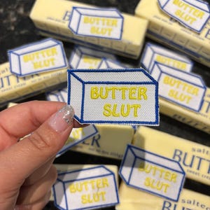 May include: A close-up of several butter sticks and embroidered patches. The patches are white with a blue outline and yellow text that reads "BUTTER SLUT". The butter sticks are yellow and have text on the packaging.