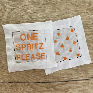 Peut inclure: Deux serviettes de cocktail en lin blanc avec des broderies orange. Une serviette dit "ONE SPRITZ PLEASE" et l'autre a un motif de petites tranches d'orange.