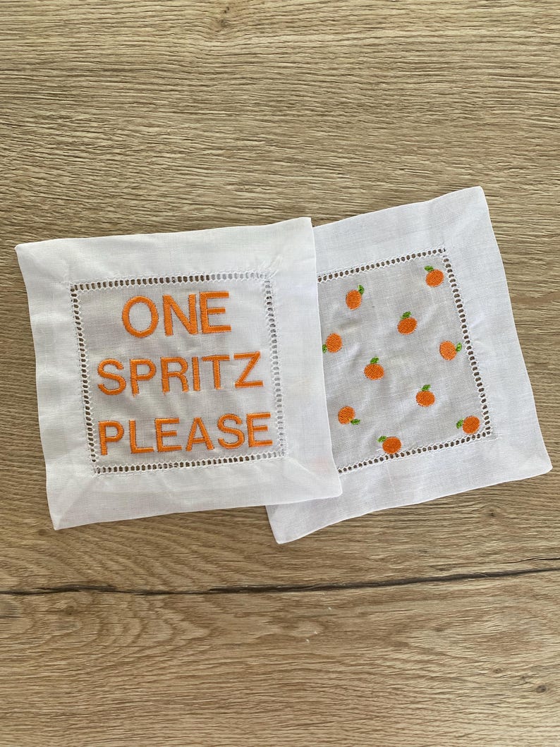 Peut inclure: Deux serviettes de cocktail en lin blanc avec des broderies orange. Une serviette dit "ONE SPRITZ PLEASE" et l'autre a un motif de petites tranches d'orange.