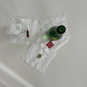 Peut inclure: Quatre serviettes en lin blanc avec des motifs brod&eacute;s. Une serviette a un motif vert et marron d'une petite bo&icirc;te, une autre a un motif rouge et vert d'une brochette avec trois objets ronds, et la troisi&egrave;me a le texte "UNIQLO" en rouge. Un verre transparent avec de la glace et une bouteille en verre vert sont pos&eacute;s sur les serviettes.
