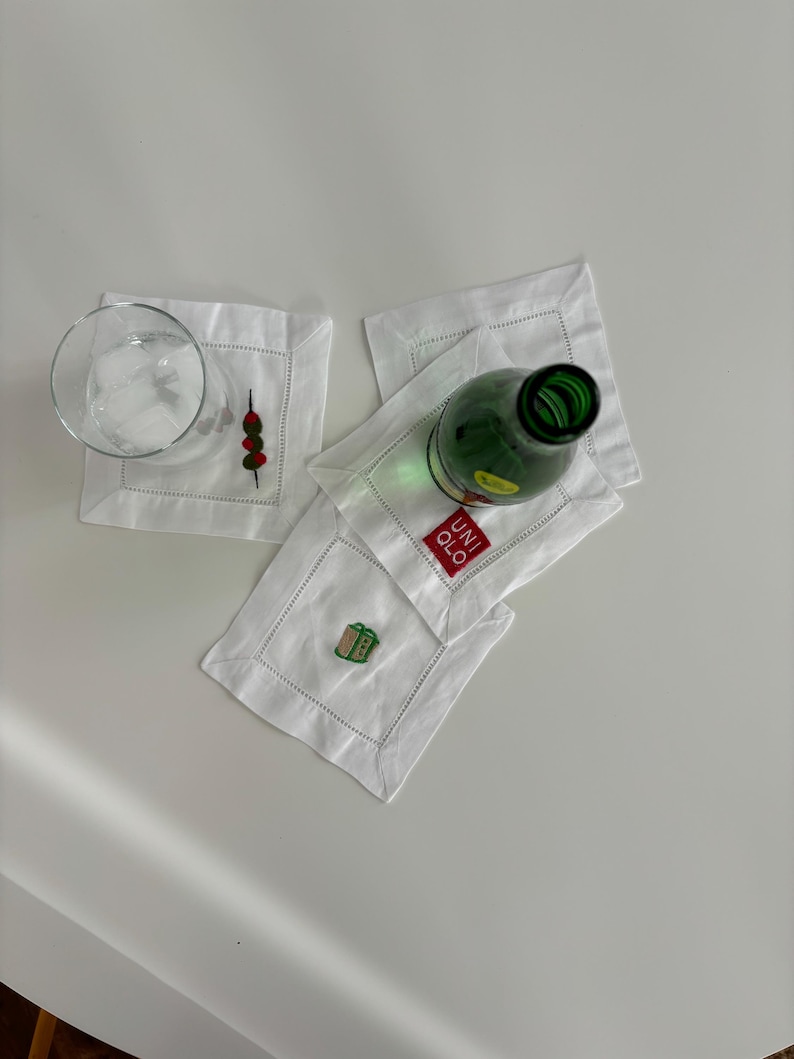 Peut inclure: Quatre serviettes en lin blanc avec des motifs brod&eacute;s. Une serviette a un motif vert et marron d'une petite bo&icirc;te, une autre a un motif rouge et vert d'une brochette avec trois objets ronds, et la troisi&egrave;me a le texte "UNIQLO" en rouge. Un verre transparent avec de la glace et une bouteille en verre vert sont pos&eacute;s sur les serviettes.