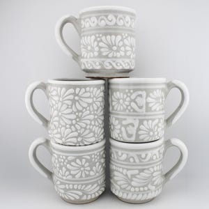 May include: A stack of five ceramic mugs featuring a light gray base color with raised white floral and geometric patterns. Each mug has a curved handle and a cylindrical shape, displaying various intricate botanical designs in relief.