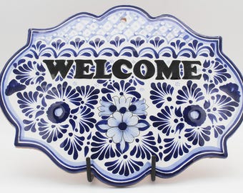 May include: A ceramic welcome sign with a white background and intricate blue floral patterns. The word "WELCOME" is prominently displayed in bold black letters. The sign has a scalloped edge and a hole for hanging.