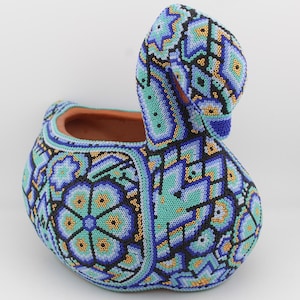 May include: A handcrafted, beaded swan-shaped decorative pot. The pot features a vibrant pattern of blue, teal, and black beads. The interior of the pot is a terracotta color. The swan's neck curves upwards, creating a unique and artistic design.