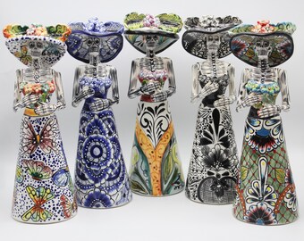 May include: Five ceramic Day of the Dead figurines, each depicting a skeleton wearing a colorful dress and a wide-brimmed hat. The figurines are all standing upright and have a variety of floral patterns.