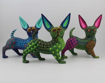 May include: Three colorful, hand-painted wooden chihuahua figurines. The dogs feature intricate patterns in blue, green, and pink, with large ears and expressive eyes. Each figurine has a unique design, showcasing the artistry of Mexican folk art.