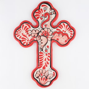 May include: A decorative wall cross featuring a cream-colored base with hand-painted red and black floral patterns. The cross has a scalloped edge outlined in bright red, with a small circular hole at the top for hanging on a wall surface.