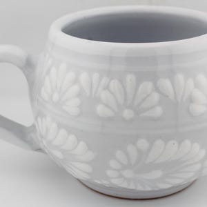 Op de afbeelding: Een ronde keramische mok met een lichtgrijze glazuurlaag en een wit reli&euml;f bloemenpatroon met herhalende bloemblaadjesmotieven in horizontale banden. De mok heeft een gebogen handvat aan de linkerkant en een gladde, ronde vorm.