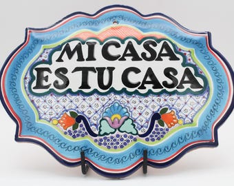 May include: A decorative ceramic plaque with a light blue border and the text "MI CASA ES TU CASA" in black. The plaque features colorful floral and geometric patterns, with a central design of a blue flower.
