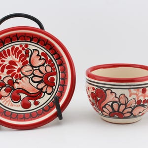 May include: A red and white ceramic teacup and saucer set with a floral design. The cup has a handle and the saucer has a black metal stand.