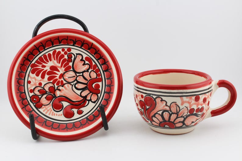 May include: A red and white ceramic teacup and saucer set with a floral design. The cup has a handle and the saucer has a black metal stand.