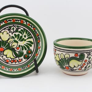May include: A green and white ceramic teacup and saucer set with a floral design. The cup has a handle and the saucer has a black metal stand.
