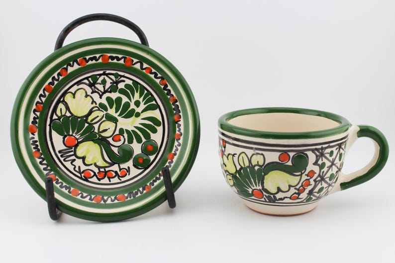 May include: A green and white ceramic teacup and saucer set with a floral design. The cup has a handle and the saucer has a black metal stand.