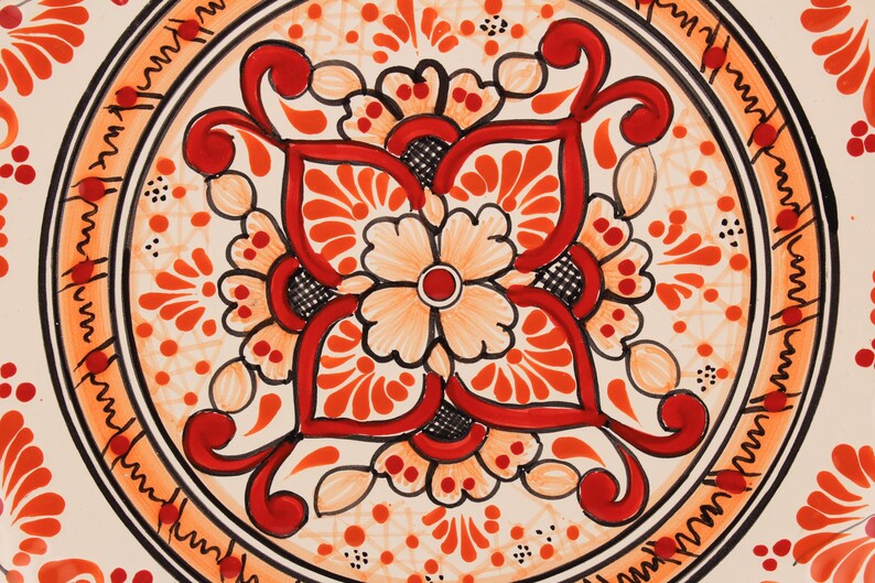 Pode incluir: Um prato de cer&acirc;mica branco com um desenho floral vermelho e laranja. O desenho apresenta uma flor central com quatro p&eacute;talos e um c&iacute;rculo menor no centro. O prato &eacute; decorado com uma borda de espirais vermelhas e laranja.