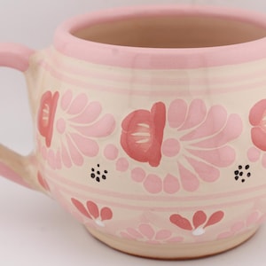 May include: A ceramic mug with a pink handle and rim, adorned with a floral pattern in shades of pink and red. The mug has a beige base color and is decorated with stylized flowers and small black dots. The mug is designed for hot beverages.